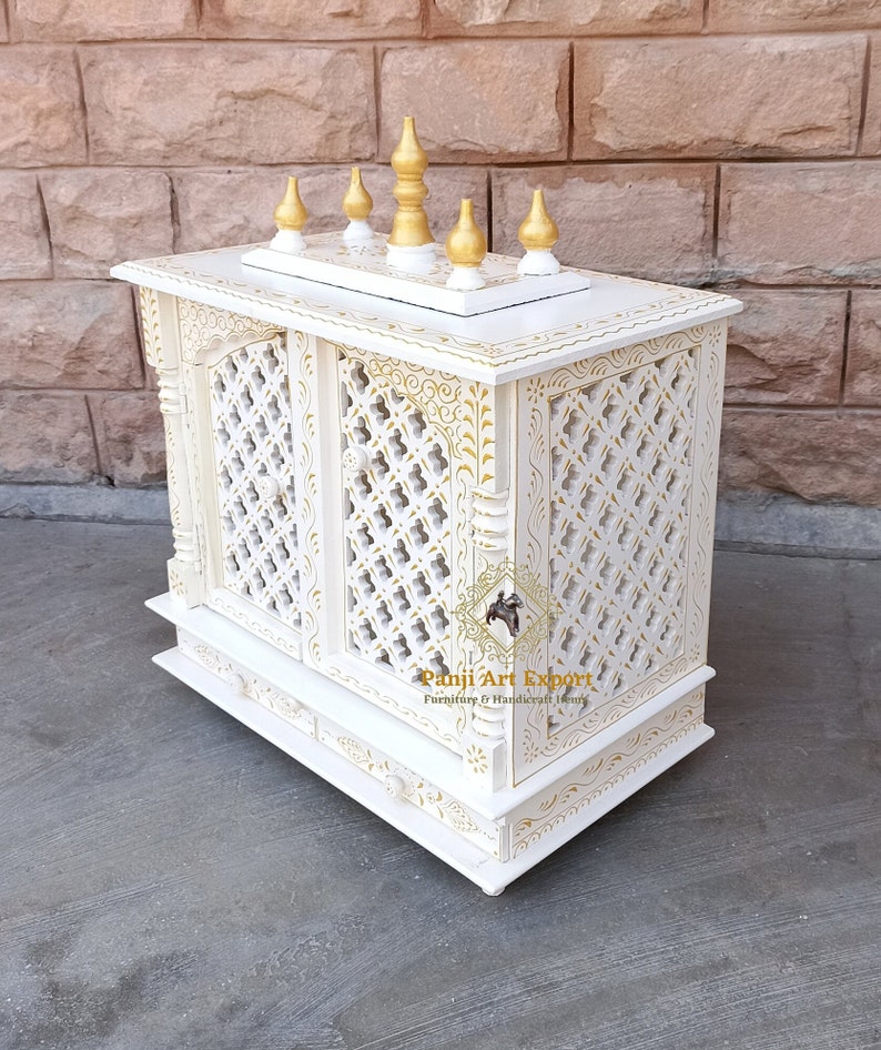 Puede incluir: Templo de madera blanco con intrincados tallados dorados y una parte superior decorativa con cinco remates dorados. El templo tiene dos puertas con un dise&ntilde;o de celos&iacute;a y un peque&ntilde;o caj&oacute;n debajo. El templo est&aacute; sentado sobre una superficie blanca.