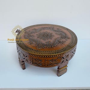 May include: A round, brown wooden table with intricate floral carvings on the top and sides. The table has short, decorative legs. The top features a central floral design surrounded by detailed leaf patterns. The table is a warm brown color.