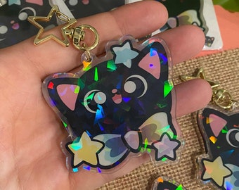 May include: A black cat keychain with iridescent glitter and stars. The keychain is made of clear acrylic and has a gold metal ring and star charm.