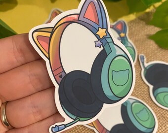 May include: A sticker of a colorful cat-eared headset with a green and blue design. The headset has a rainbow headband and a star on the side. The sticker is on a hand.