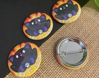 May include: Four round pinback buttons with a black cat design. The cat is wearing a purple bow and has a skull on its chest. The buttons have a yellow background with orange flames and the text "Let it Burn". One button is missing the pinback.
