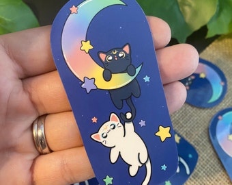 May include: A sticker featuring two cartoon cats on a crescent moon with a rainbow gradient. The black cat is hanging from the moon, while the white cat is reaching up to grab the black cat's tail. The background is a dark blue with white stars.