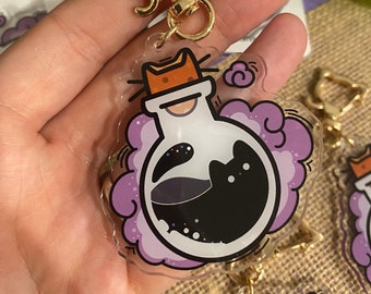 May include: A purple and black cat charm keychain with a gold cat head clasp. The charm features a black cat inside a clear glass bottle with a purple cloud around it.