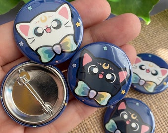 May include: Four 1-inch button pins featuring a black cat and a white cat with a crescent moon and stars on a dark blue background. The cats are wearing a rainbow bow tie.