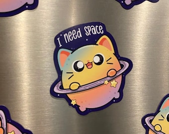 Può includere: Quattro magneti a forma di pianeta gatto colorati con il testo "Ho bisogno di spazio" su due di essi. I gatti sono gialli, blu e rosa con un anello viola intorno. I magneti sono su una superficie argentata.