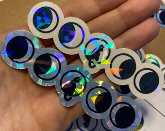 Può includere: Un set di adesivi olografici iridescenti con un design di gatto nero e luna crescente. Gli adesivi sono rotondi e hanno uno sfondo bianco con un contorno nero.