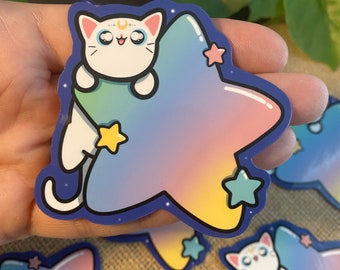 May include: A white cat with a crescent moon on its forehead sits on a rainbow star with yellow stars. The sticker is on a brown surface.