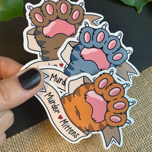 May include: Three cat paw stickers in brown, blue, and orange, each with a pink paw pad and claws. A banner on each sticker reads "Murder Mittens" in black text with a red heart. The stickers are on a neutral background.