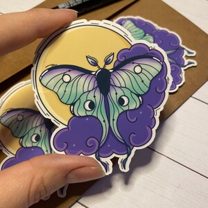 May include: A sticker featuring a green and purple moth with large eyes, set against a yellow moon and purple clouds.
