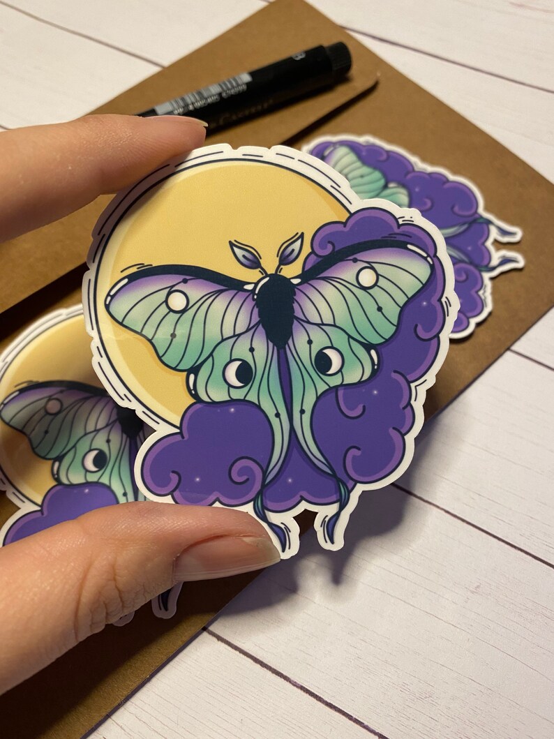 May include: A sticker featuring a green and purple moth with large eyes, set against a yellow moon and purple clouds.