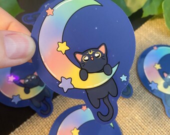 May include: A black cat with a crescent moon on its forehead hangs from a crescent moon with a rainbow gradient. The moon is surrounded by stars on a dark blue background.
