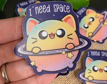 May include: A sticker featuring a cartoon cat with a rainbow gradient, holding a planet with a ring around it. The cat has a happy expression and the text "I need space" is written above it.
