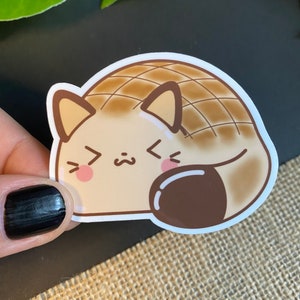 May include: A brown and tan cartoon cat sticker with a brown swirl design on its back. The cat has a cute expression with closed eyes and a small mouth. The sticker is on a brown and white textured surface.