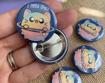 May include: Four colorful cat-themed button pins with the text "I need space" on the front. The pins are on a brown surface.