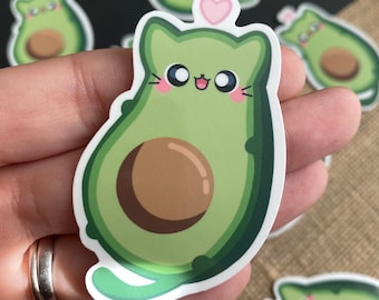 May include: A green avocado sticker with a cute cartoon cat face and a pink heart. The cat is wearing a green avocado costume and has a brown seed in the middle of its body.