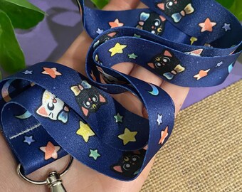 May include: A blue lanyard with a white cat and black cat design. The lanyard has a silver clip and features stars and crescent moons.