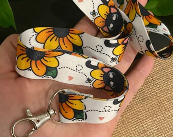 May include: White lanyard with a black cat design inside a yellow sunflower. The lanyard has a silver metal clip.