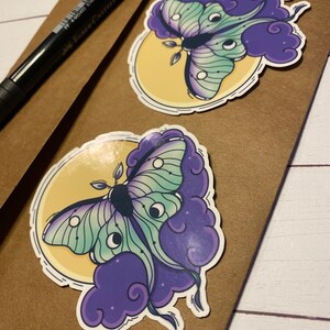 May include: Two stickers featuring a green and purple moth with large eyes on a yellow moon and purple clouds.