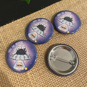 May include: Three black cat UFO pins with the text "Feed me, human!" on a purple background with stars. The back of one pin is shown, revealing a silver metal pin back.