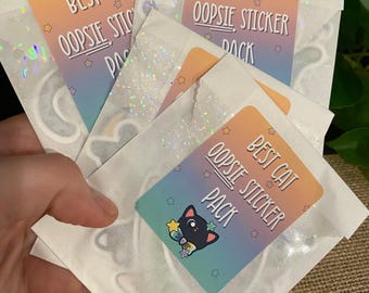 May include: A set of four white envelopes with holographic glitter. Each envelope has a sticker with the text "Best Cat Oopsie Sticker Pack".