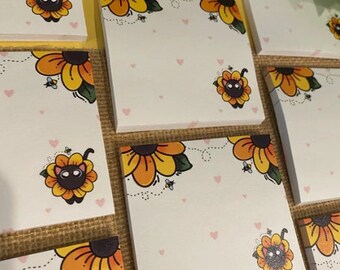 May include: White square notepads with a sunflower and cat design. The sunflowers are yellow and orange with green leaves. A cartoon cat is in the center of the sunflower. Small pink hearts and bees are also on the notepads.