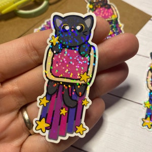 Puede incluir: Un gato negro con un disfraz de tostada brillante rosa y blanco con un rastro de estrellas arcoíris detrás. La pegatina es holográfica y tiene un acabado brillante.