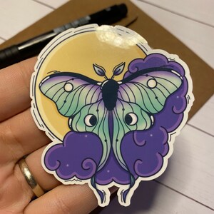 May include: A sticker featuring a green and purple luna moth with a yellow moon and purple clouds in the background.