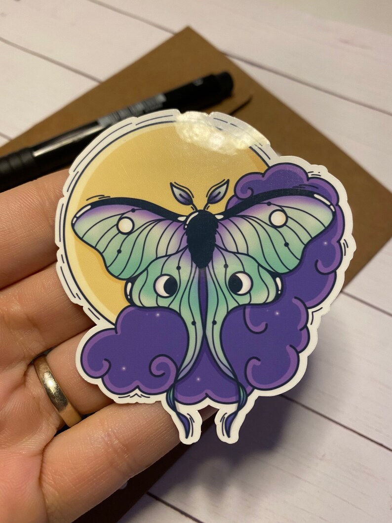 May include: A sticker featuring a green and purple luna moth with a yellow moon and purple clouds in the background.