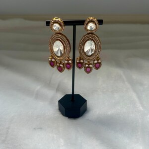 May include: Gold-toned chandelier earrings with clear and ruby-colored stones. The earrings feature a large oval center stone, surrounded by smaller stones, and ruby-colored teardrop dangles. The earrings are displayed on a black stand.
