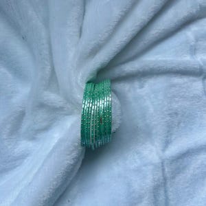 May include: A stack of light green bangles with small, evenly spaced dots. The bangles are resting on a soft, white, textured surface. The bangles are a solid color and appear to be made of metal or glass.