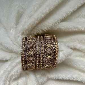 May include: A set of four gold-toned metal bangles featuring an intricate embossed pattern with small white faux pearls embedded in oval-shaped settings. The bangles have a textured, antique-style finish with delicate beaded edges along the top and bottom.