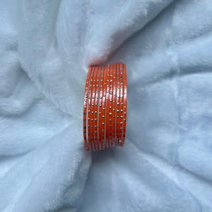 May include: A stack of orange bangles with small silver dots. The bangles are stacked together, and the color is a vibrant orange. The background is a soft, white, fluffy material.