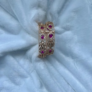 May include: Two gold-toned hinged bangles featuring a repeating pattern of teardrop-shaped fuchsia stones and small clear crystals arranged in floral motifs. The metalwork includes intricate filigree detailing along the edges of the bracelets.