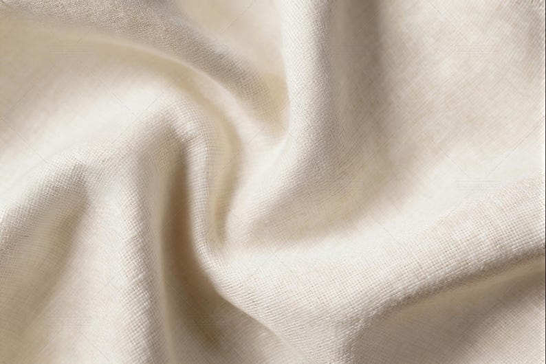 Puede incluir: Una pieza de tela color blanco roto con una textura tejida visible y un aspecto natural sin blanquear. El material est&aacute; drapeado en pliegues suaves y fluidos que crean sombras y reflejos sutiles en la superficie del tejido.