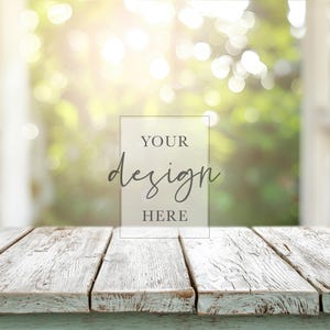 Puede incluir: Una mesa rústica de madera pintada de blanco con un aspecto envejecido. La mesa está frente a un fondo borroso de una ventana y vegetación exuberante. El texto "YOUR design HERE" se muestra en un rectángulo transparente.