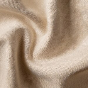 Puede incluir: Una pieza de tela de color beige claro con una textura tejida visible y un acabado mate suave. El material est&aacute; drapeado para crear pliegues profundos y fluidos, con sombras que resaltan el grano natural y el color uniforme del tejido.
