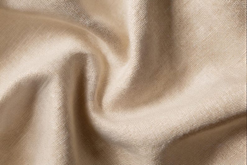 Puede incluir: Una pieza de tela de color beige claro con una textura tejida visible y un acabado mate suave. El material est&aacute; drapeado para crear pliegues profundos y fluidos, con sombras que resaltan el grano natural y el color uniforme del tejido.