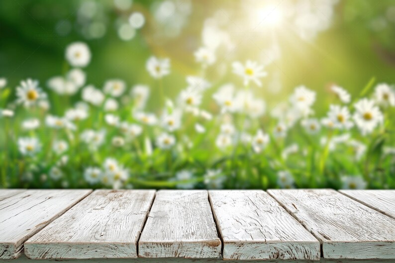 Puede incluir: Una superficie de mesa de madera blanca desgastada con vetas y textura visibles, compuesta por tablones horizontales unidos, situada frente a un campo desenfocado de margaritas blancas y hierba verde bajo luz solar brillante.