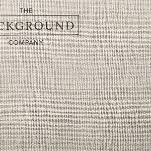 May include: A light beige fabric with a visible woven texture. In the upper left corner, a black rectangular border encloses the text THE BACKGROUND COMPANY, with the word BACKGROUND inside the box and THE and COMPANY placed above and below.