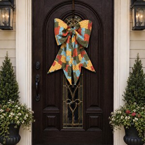 May include: A large decorative bow made of fabric with a patchwork pattern in shades of yellow, red, and green. The bow features two prominent loops at the top and two long, pointed tails hanging downward, centered on a dark brown wooden door.