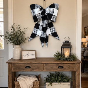 May include: A large decorative bow featuring a black and white buffalo check pattern. The bow is constructed from fabric with wide loops and long, pointed tails hanging downward, suspended from a black wall hook by a thin, neutral-colored cord.