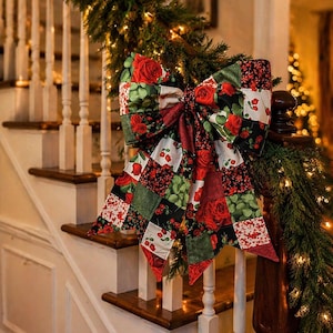 May include: A large decorative bow made from a patchwork fabric featuring a pattern of red roses, green leaves, and black and white floral squares. The bow has two long, pointed tails and is attached to a green garland with warm white lights.