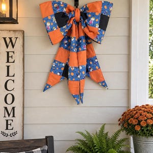 May include: A large decorative bow made of fabric featuring a patchwork design with orange, black, and blue sections. The blue fabric displays a repeating pattern of cartoon characters and pumpkins. The bow has two long, pointed tails hanging down.