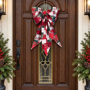 May include: A large decorative bow made of fabric with a patchwork pattern featuring red, green, white, and black plaid designs. The bow has two wide loops and two long, pointed tails hanging downwards, suspended by a gold hook on a dark wood door.