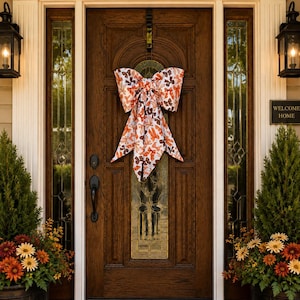 May include: A large decorative bow featuring a white fabric base with a repeating pattern of orange and brown butterflies. The bow is hung on a dark brown wooden door with a glass panel insert, positioned centrally above the door handle hardware.