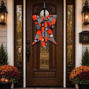 May include: A large fabric bow hanging on a dark brown wooden door. The bow features a pattern of black and white buffalo check squares alternating with solid orange sections and small pumpkin illustrations in orange, white, and black colors.