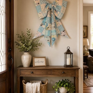 May include: A welcoming entryway featuring a wooden console table with a decorative bow, a framed "welcome" sign, and a lantern. The bow is made of patchwork fabric in shades of blue and white. A basket with a blanket and a vase with greenery add to the decor.