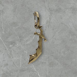 May include: A single gold-colored earring with a geometric sword design. The earring features a hoop at the top and a stylized sword pendant with sharp angles and a pointed tip. The earring is displayed against a gray marble background.