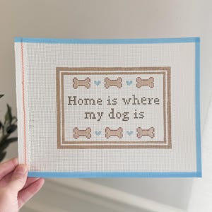 May include: A needlepoint canvas with a light blue border and the text "Home is where my dog is." The design includes brown dog bone shapes and blue hearts. The canvas is white with a grid pattern.