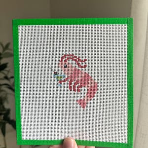 May include: A cross-stitch design featuring a pink shrimp holding a small object, set against a white background. The design is framed by a green border. The shrimp is detailed with darker pink accents.